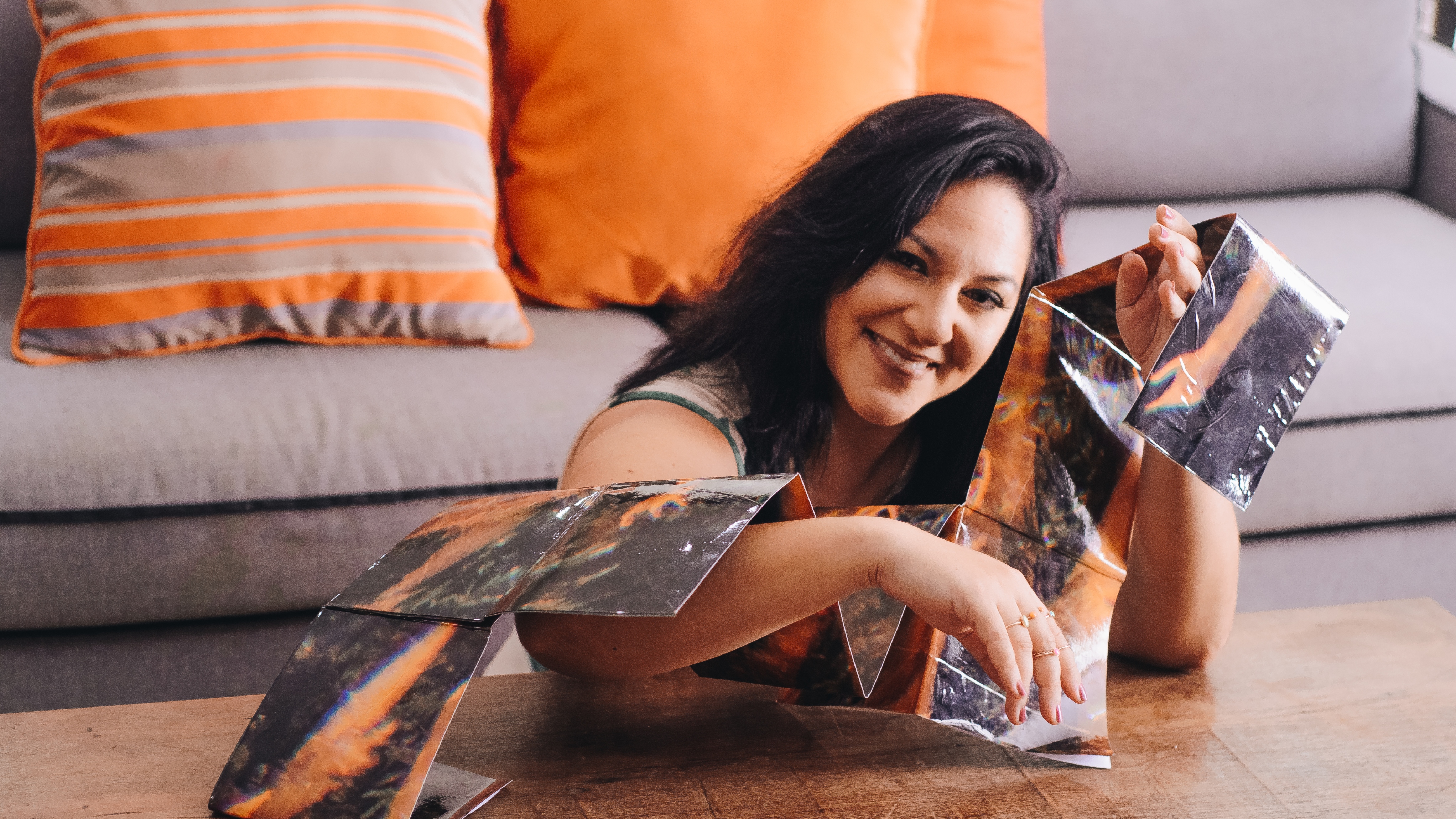 A artista Ana Mundim, mulher branca de cabelos longos e pretos, está sentada atrás de uma mesa de madeira, em uma sala com sofá ao fundo. Ela sorri para a câmera enquanto segura e veste uma estrutura de papelão ou material semelhante, com partes refletivas e coloridas. Ao fundo, há almofadas em tons de laranja e cinza.